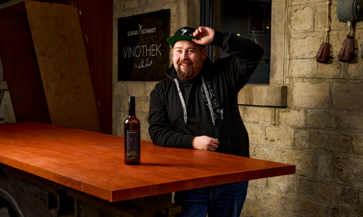 Mann in schwarzem Hoodie und Kappe steht lächelnd hinter rustikalem Holztisch – lehnt sich mit einer Hand auf die Tischplatte, die andere am Mützenschirm, vor ihm eine Weinflasche, Hintergrund mit heller Steinwand, Fenster, Laterne und Schild „LUKAS SCHMIDT VINOTHEK & BAR“, Szene vermittelt entspannte Atmosphäre und persönlichen Bezug zur Weinbar.