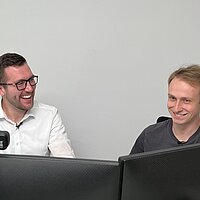 Zwei Personen sitzen nebeneinander vor Computerbildschirmen, lächeln und interagieren – eine Webcam ist montiert.
