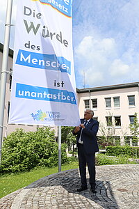 Person steht auf einem gepflasterten Platz vor einem Gebäude und befestigt ein großes Banner an einem Fahnenmast mit der Aufschrift „Die Würde des Menschen ist unantastbar“ sowie dem Logo „Frankens MEHR Region“.
