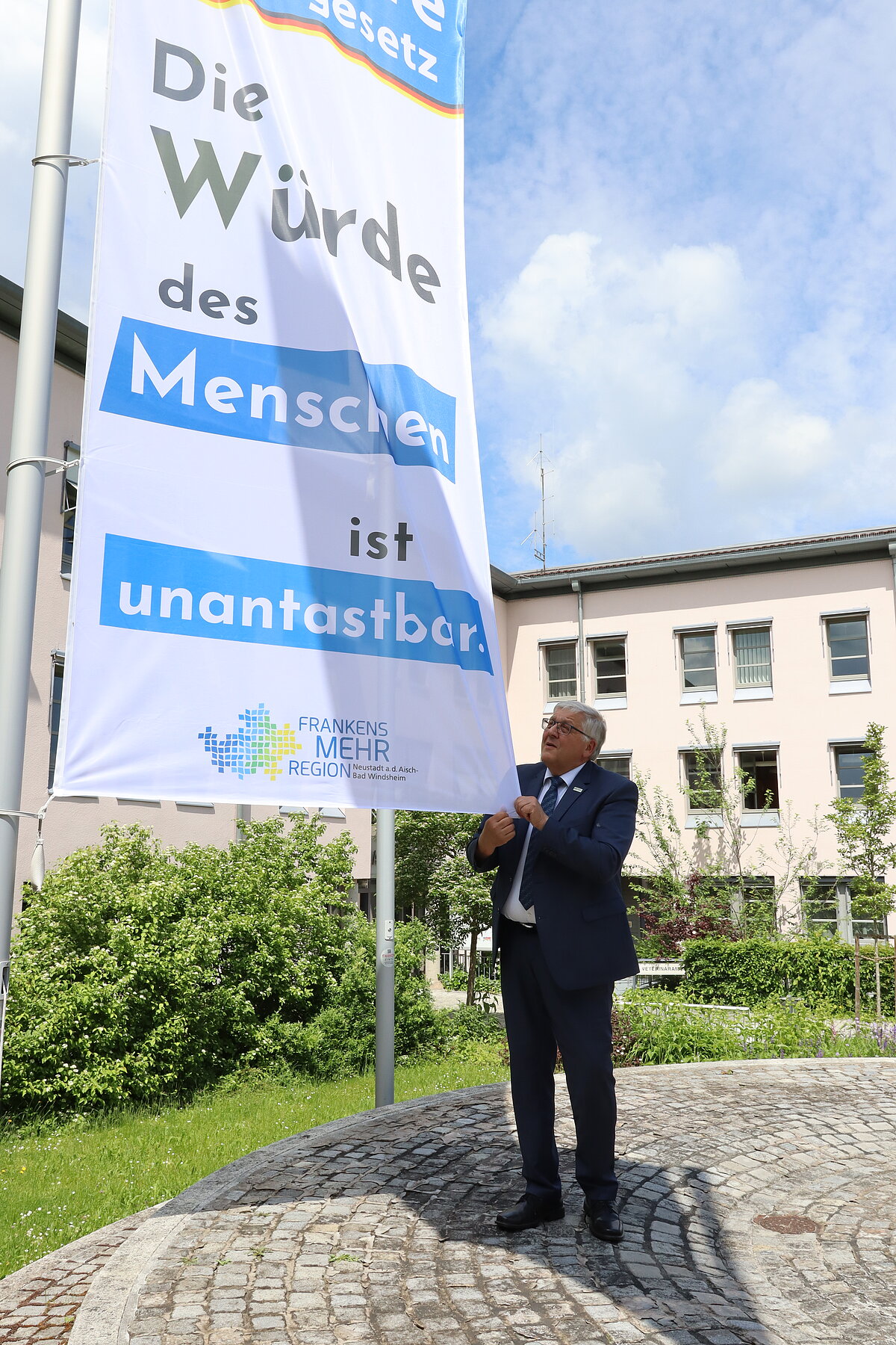 Person steht auf einem gepflasterten Platz vor einem Gebäude und befestigt ein großes Banner an einem Fahnenmast mit der Aufschrift „Die Würde des Menschen ist unantastbar“ sowie dem Logo „Frankens MEHR Region“.