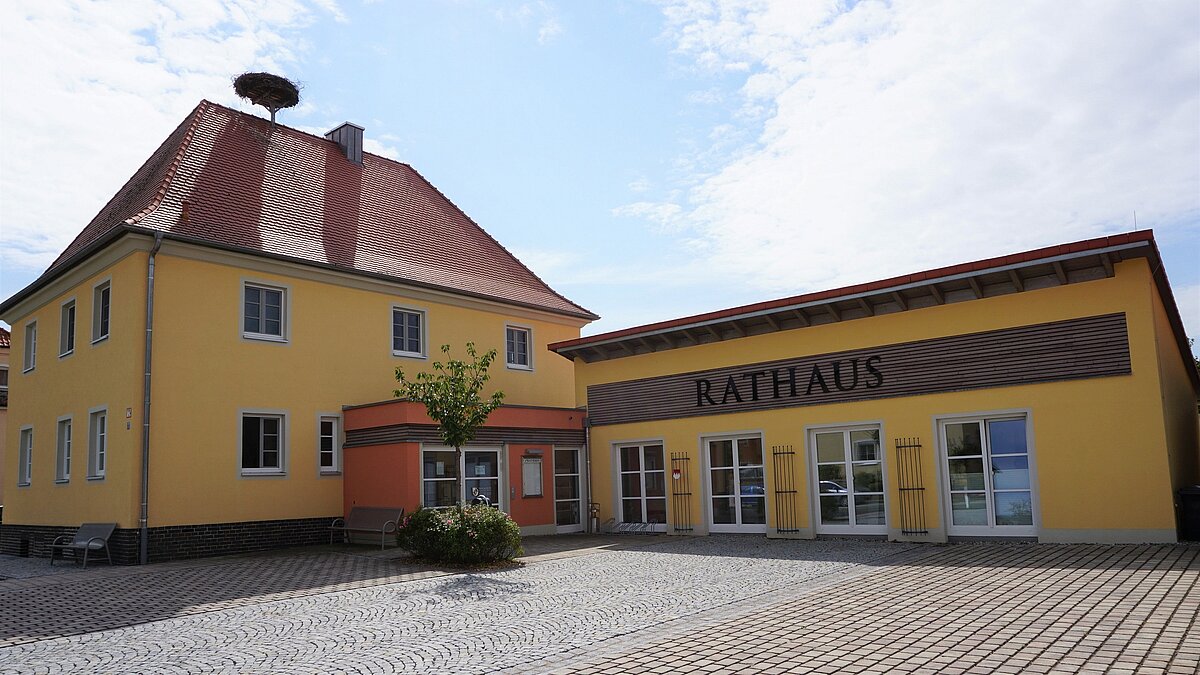 Zwei Gebäude auf einem gepflasterten Platz: links ein zweistöckiges Haus mit gelber Fassade, rotem Ziegeldach und einem Storchennest auf dem Dach; rechts ein eingeschossiges Gebäude mit der Aufschrift „RATHAUS“. Beide Gebäude haben Eingangsbereiche mit farblichen Akzenten. Vor dem Rathaus stehen Bänke und ein kleiner Baum.