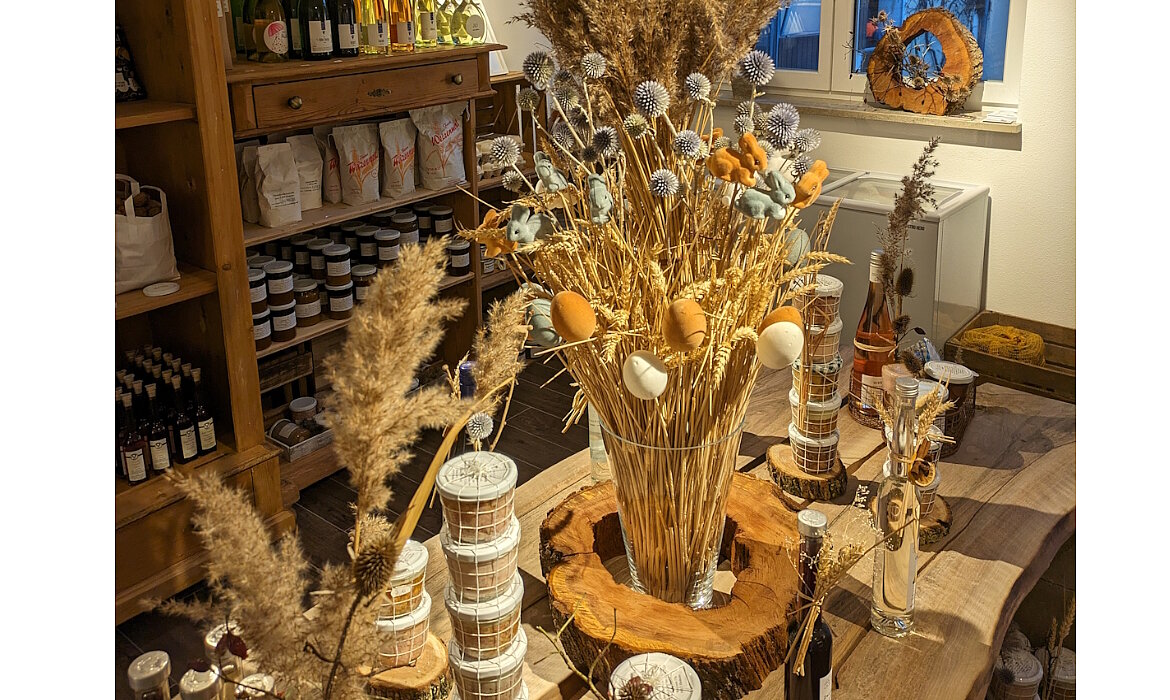Ein liebevoll dekorierter Tisch mit Trockenblumen, bunten Eiern und kleinen Tierfiguren – umgeben von Gläsern, Körben und handgemachten Produkten. Dieses Arrangement strahlt ländliche Gemütlichkeit und saisonale Festlichkeit aus – ein echter Blickfang für Hofladen oder Zuhause.