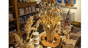 Ein liebevoll dekorierter Tisch mit Trockenblumen, bunten Eiern und kleinen Tierfiguren – umgeben von Gläsern, Körben und handgemachten Produkten. Dieses Arrangement strahlt ländliche Gemütlichkeit und saisonale Festlichkeit aus – ein echter Blickfang für Hofladen oder Zuhause.