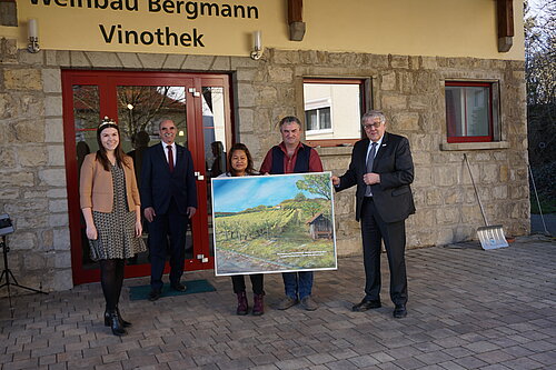 Fünf Personen vor dem Gebäude „Weinbau Bergmann Vinothek“ – zwei halten ein Gemälde mit Weinlandschaft, Szene deutet auf kulturelles Ereignis oder Präsentation im Kontext regionaler Weinkultur.