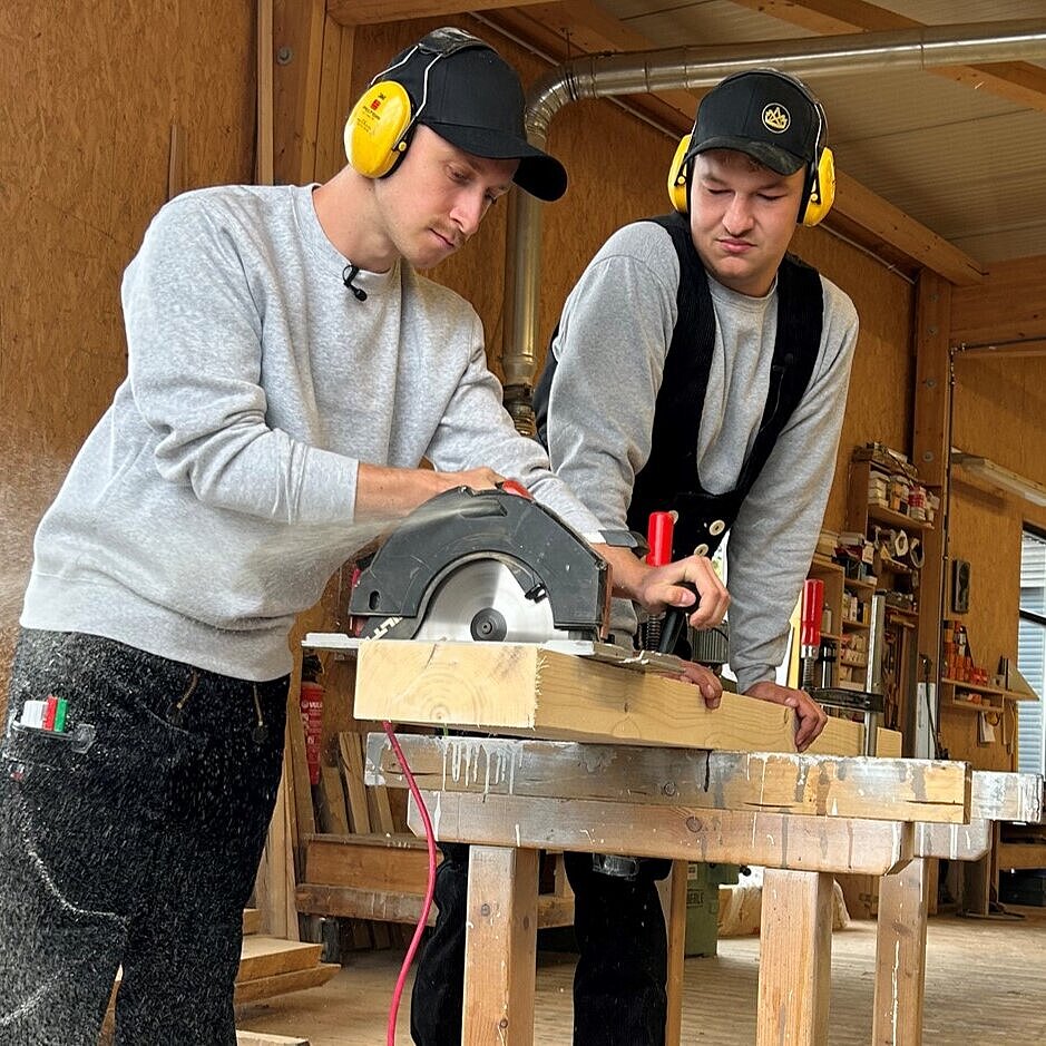 Zwei Personen arbeiten in einer hellen Holzwerkstatt mit Gehörschutz und Mikrofon-Headset an einer Kreissäge – deutet auf eine praktische Schulung oder berufliche Anleitung im Rahmen der „JOB JUNGLE“-Initiative.