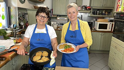 Zwei Frauen stehen in einer Küche mit grünen Schränken. Beide tragen blaue Schürzen. Eine Person hält eine Pfanne mit vier goldbraunen Bratlingen, die andere präsentiert einen Teller mit ähnlichen Bratlingen und gegartem Gemüse wie Karotten und Bohnen. Die Szene wirkt wie eine Kochvorführung.