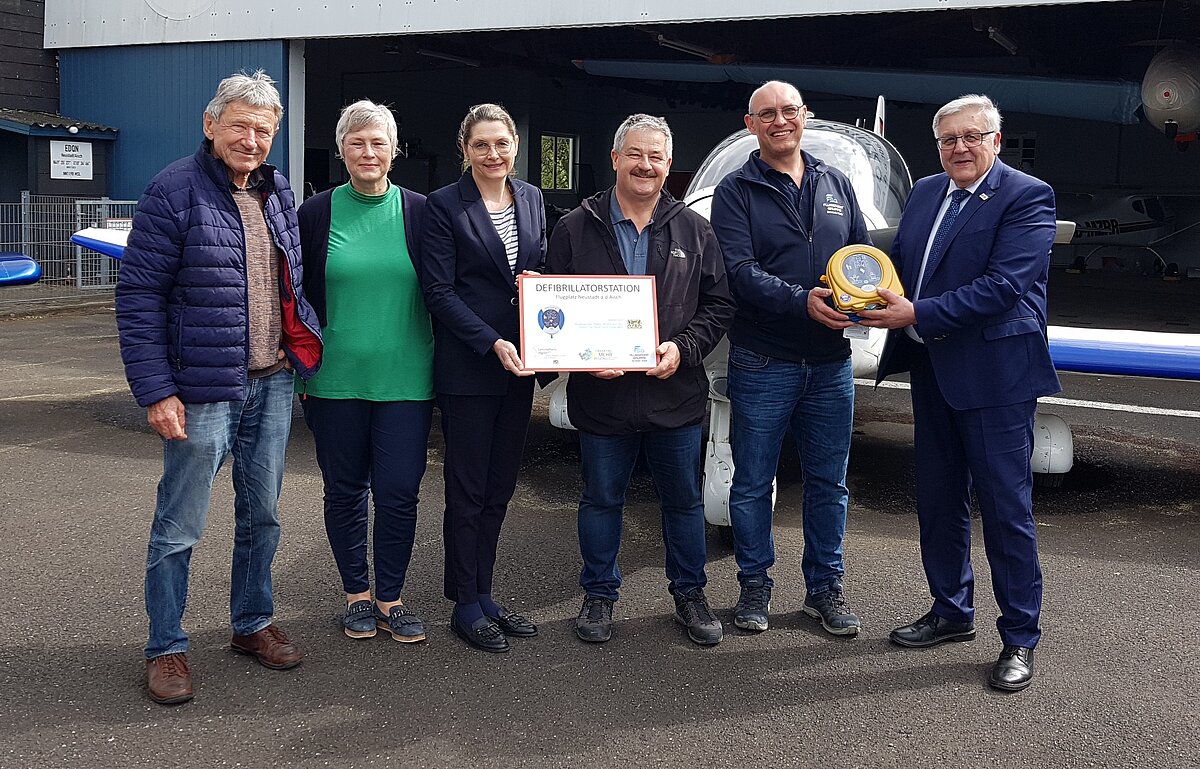 Personen vor Flugzeug mit Überreichung Defibrillator
