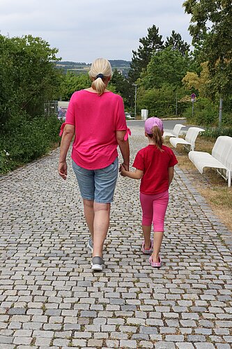 Eine erwachsene Person in pinkem Oberteil und ein Kind in roter Kleidung gehen Hand in Hand auf einem gepflasterten Weg. Rechts stehen weiße Bänke, links und im Hintergrund ist grüne Vegetation zu sehen. Der Himmel ist bewölkt.