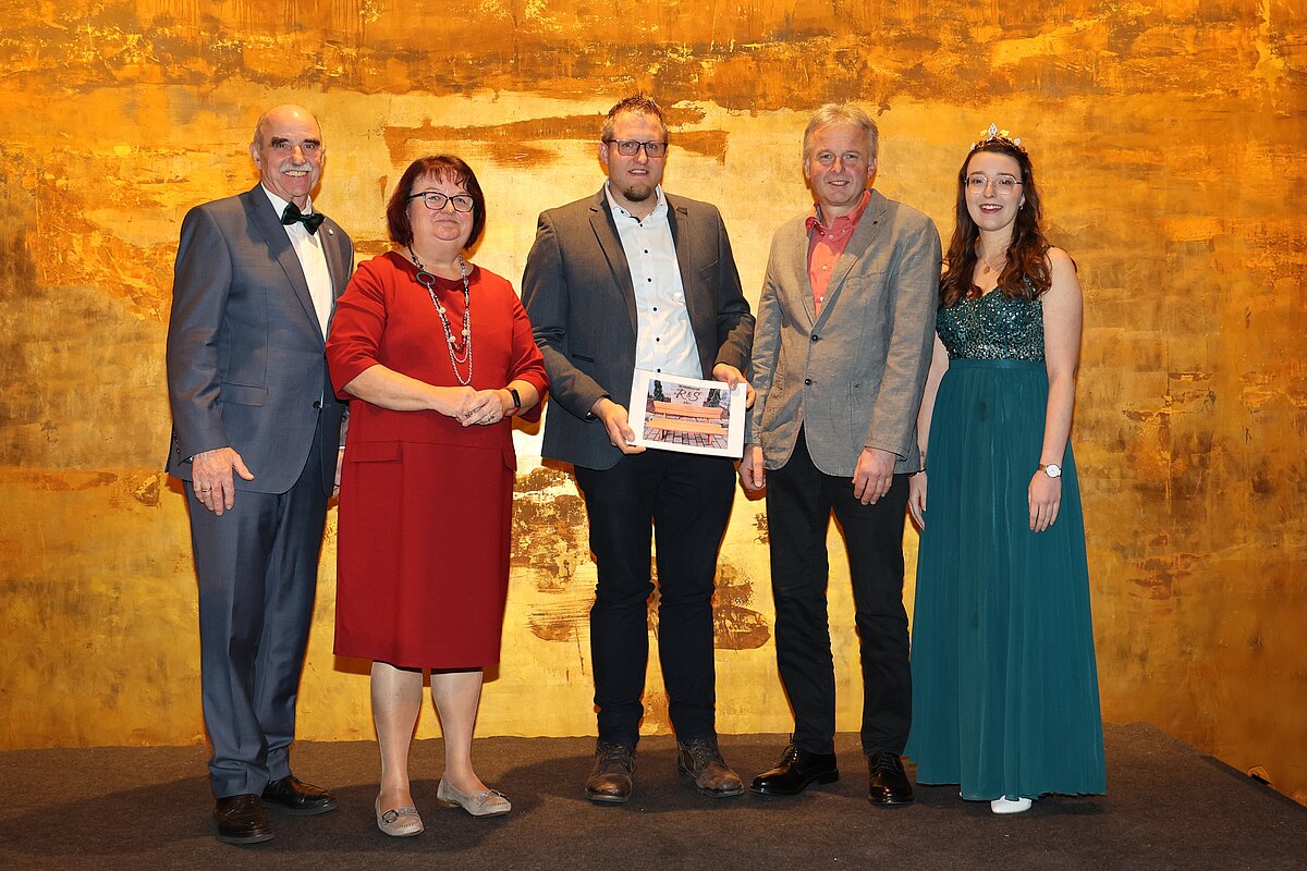 Fünf Personen stehen vor goldfarbener Bühnenkulisse, mittig wird eine gerahmte Urkunde präsentiert – formelle Kleidung und Gruppenpose deuten auf eine Auszeichnung oder Ehrung hin.