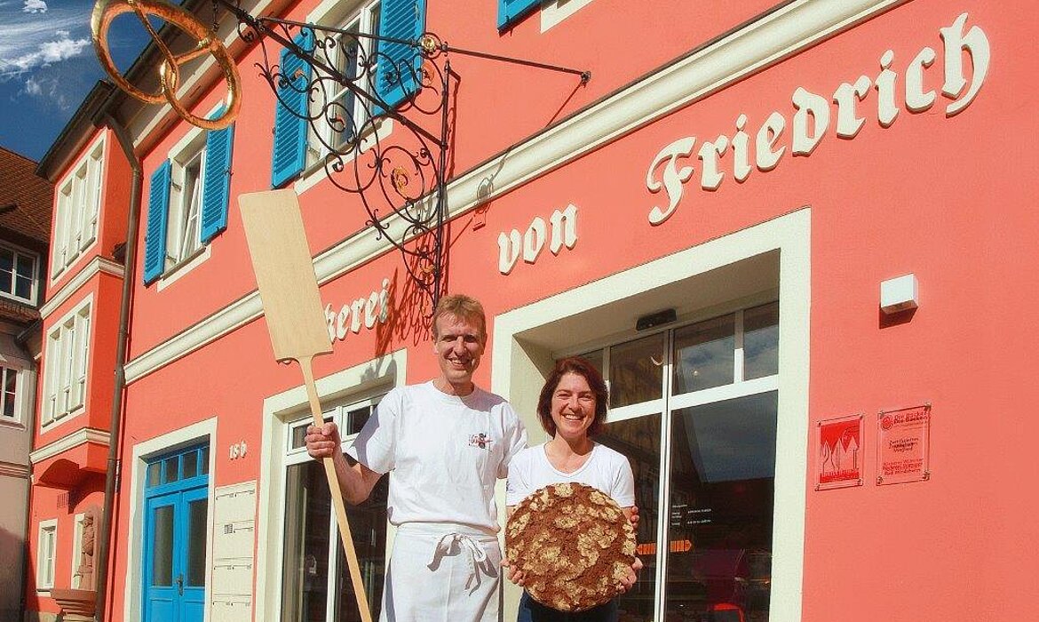 Vor der farbenfrohen „Bäckerei von Friedrich“ präsentieren zwei Bäcker stolz ihr Handwerk – einladende Szene, die Tradition und Genuss vereint.