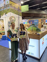 Zwei Personen in festlicher Tracht vor Messestand der Hochschulen Würzburg-Schweinfurt und München – sie halten Infomaterial, im Hintergrund Dekoration mit großem Fisch und weiteren Ausstellern, Szene zeigt lebendige Präsentation bei Bildungs- oder Regionalveranstaltung.